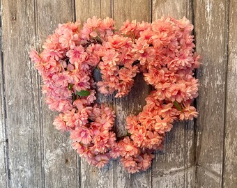 May include: A heart-shaped wreath composed of numerous small, light pink flowers. The wreath is mounted on a weathered, gray wooden door. The flowers are densely packed, creating a full, textured appearance. A few green leaves are visible.