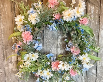 May include: A wreath made of artificial flowers, featuring pink, white, and blue blossoms. The wreath is made on a green base and is perfect for spring or summer decor.