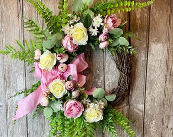 May include: A pink and white floral wreath with a pink satin bow. The wreath is made with artificial flowers and greenery, including roses, peonies, and ferns.