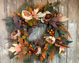 May include: A fall-themed wreath with faux peonies, eucalyptus leaves, and dried grasses in shades of brown, beige, and green. The wreath is decorated with small berries and acorns.
