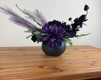 Könnte beinhalten: Ein Blumenarrangement in einer mattschwarzen, kugelförmigen Vase mit tiefvioletten Dahlienblüten, dunklen, orchideenartigen Blumen und federartigen, violett getönten Ziergräsern. Grünes, farnartiges Blattwerk akzentuiert die Basis des Gestecks.