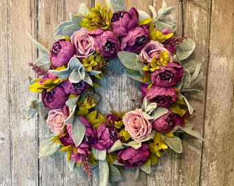 May include: A floral wreath featuring a circular arrangement of purple peonies, pink roses, and green and gray foliage. The wreath is displayed against a weathered, wooden door, creating a rustic aesthetic. The wreath is a decorative item.