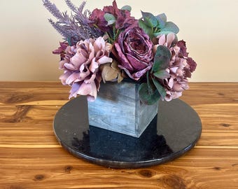 May include: A floral arrangement in shades of purple and pink, featuring peonies, roses, and greenery, is displayed in a rustic wooden box. The arrangement is placed on a black circular base.