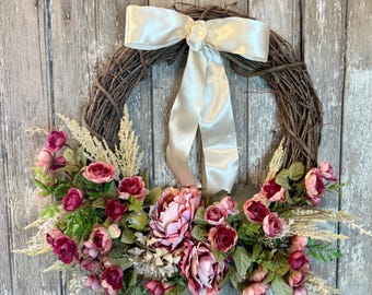 May include: A decorative wreath with a natural brown twig base, adorned with pink and burgundy flowers, green leaves, and dried wheat. A cream-colored satin bow is tied at the top. The wreath is displayed against a weathered wooden door.
