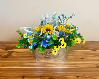 May include: A metal planter with the text "Flowers & Garden" on the front, filled with a floral arrangement of yellow sunflowers, blue and white flowers, and green foliage.