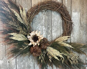 May include: A rustic grapevine wreath decorated with dried grasses, a faux sunflower, and brown pinecones. The wreath is a natural, earthy color and would be a beautiful addition to any home decor.