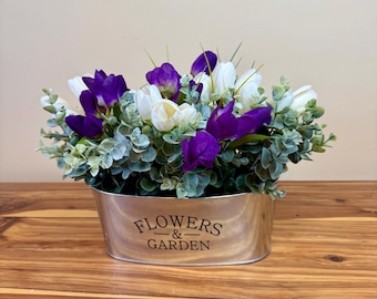 May include: A decorative arrangement of purple and white flowers with green foliage in a metallic oval container. The container has the words "FLOWERS & GARDEN" printed on the front. The arrangement sits on a wooden surface.