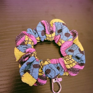 May include: A colorful scrunchie with a black and white pattern, featuring a pink, yellow, and blue color scheme. It has a silver metal clip attached.