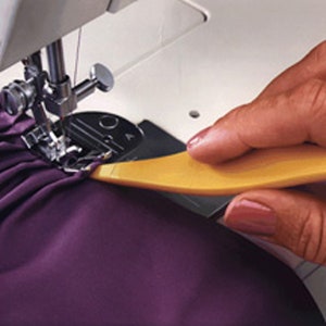 May include: A yellow plastic seam ripper tool being used to remove excess fabric from a purple garment while being sewn on a sewing machine.