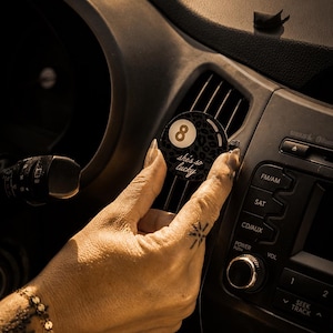 May include: A circular car air freshener vent clip featuring a black and grey leopard print pattern, a white circle with the number 8 in the center, and the cursive text she's so lucky printed in white below the number on the black surface.