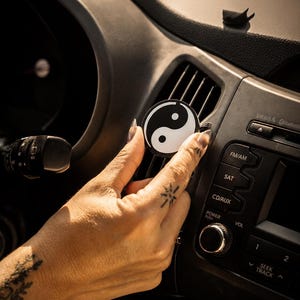 May include: A circular car air freshener clip featuring a black and white yin yang symbol. The round accessory is designed to attach to the horizontal slats of a vehicle's dashboard air vent, providing a contrasting monochromatic graphic design.