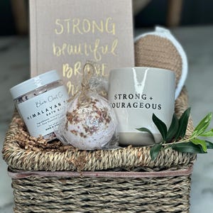 Könnte beinhalten: Ein Weidenkorb gefüllt mit Selbstpflegeartikeln, darunter eine Badebombe, eine Tasse mit der Aufschrift "Strong + Courageous", ein Buch mit der Aufschrift "Strong beautiful brave" und ein kleines Glas mit Himalaya-Badesalz.