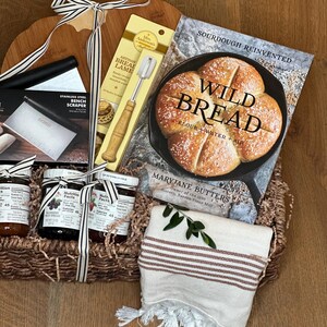 May include: A bread-making gift basket with a wooden cutting board, a stainless steel bench scraper, a bread lame, a cookbook titled "Wild Bread", and jars of fruit spread. A striped towel and decorative ribbon complete the arrangement.