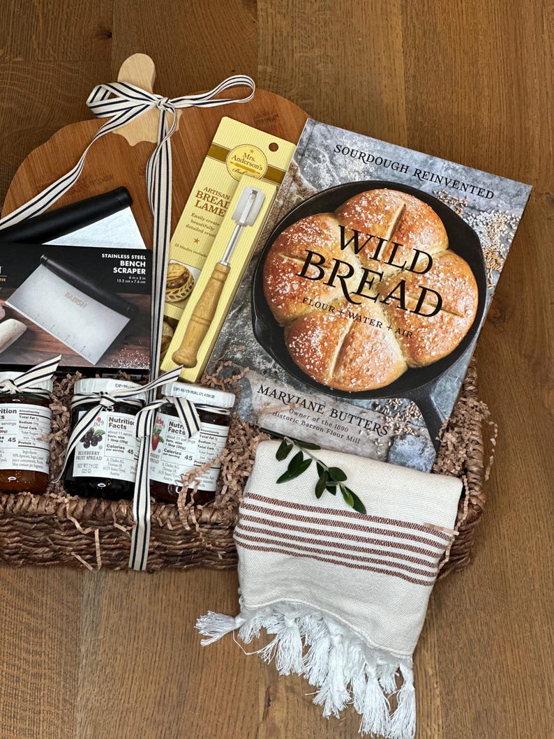 May include: A bread-making gift basket with a wooden cutting board, a stainless steel bench scraper, a bread lame, a cookbook titled "Wild Bread", and jars of fruit spread. A striped towel and decorative ribbon complete the arrangement.