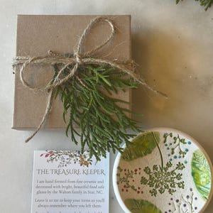 May include: A small, white ceramic dish with green and blue floral designs. The dish is hand-painted and has a rustic, handmade look. The dish is sitting on a white surface. The text on the card reads: "THE TREASURE KEEPER I am hand formed from fine ceramic and decorated with bright, beautiful food safe glazes by the Walton family in Star, NC. Leave it to me to keep your items so you'll always remember where you left them. From clay to glazes to the box that holds me, I'm locally sourced and ready to keep your small treasures at hand."