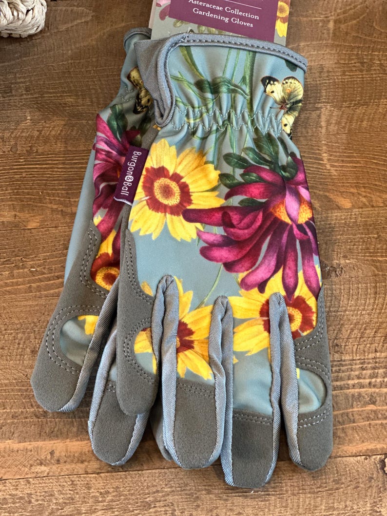 May include: A pair of gray gardening gloves with a floral print featuring yellow, red, and white flowers. The gloves have a  Burgon & Ball logo on the wrist.