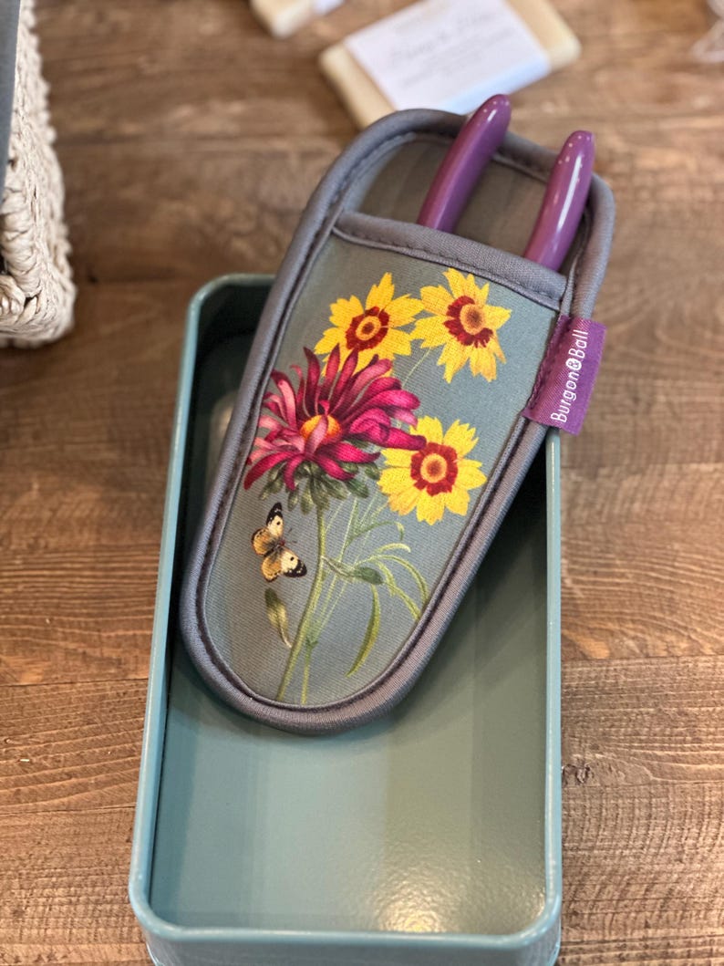 May include: A gray gardening tool holder with a floral print featuring yellow sunflowers, pink flowers, and a butterfly. The holder has two purple gardening shears inside and the text "Burgoyne & Ball" on the side.