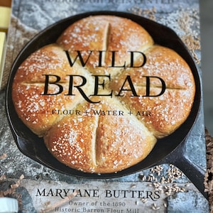 May include: A cookbook titled "Wild Bread" featuring a close-up of a freshly baked loaf of bread in a cast iron skillet. The cover text includes "Flour + Water + Air" and the author's name, Mary Jane Butters, owner of the Historic Barron Flour Mill.