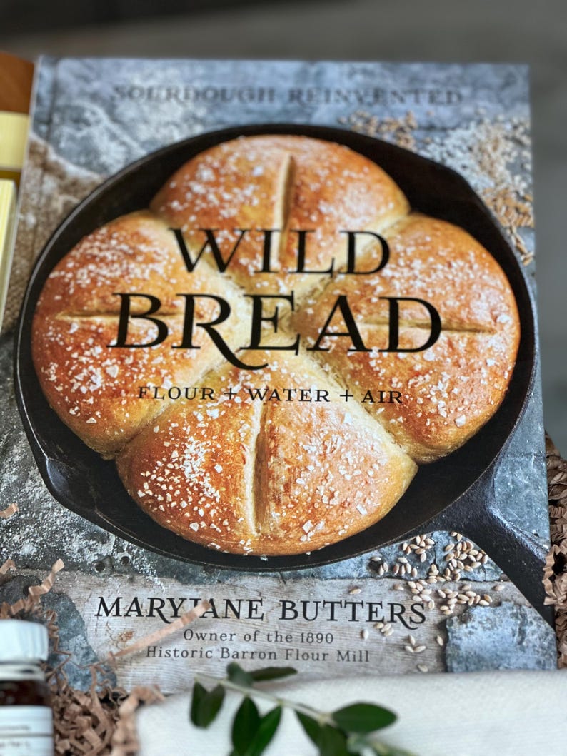 May include: A cookbook titled "Wild Bread" featuring a close-up of a freshly baked loaf of bread in a cast iron skillet. The cover text includes "Flour + Water + Air" and the author's name, Mary Jane Butters, owner of the Historic Barron Flour Mill.
