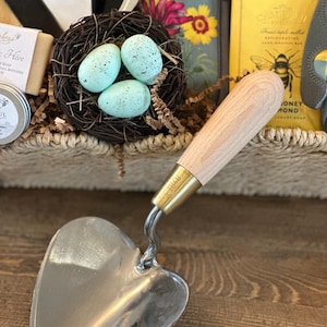 May include: A heart-shaped gardening trowel with a wooden handle and a brass ferrule. The trowel is sitting on a wooden surface.