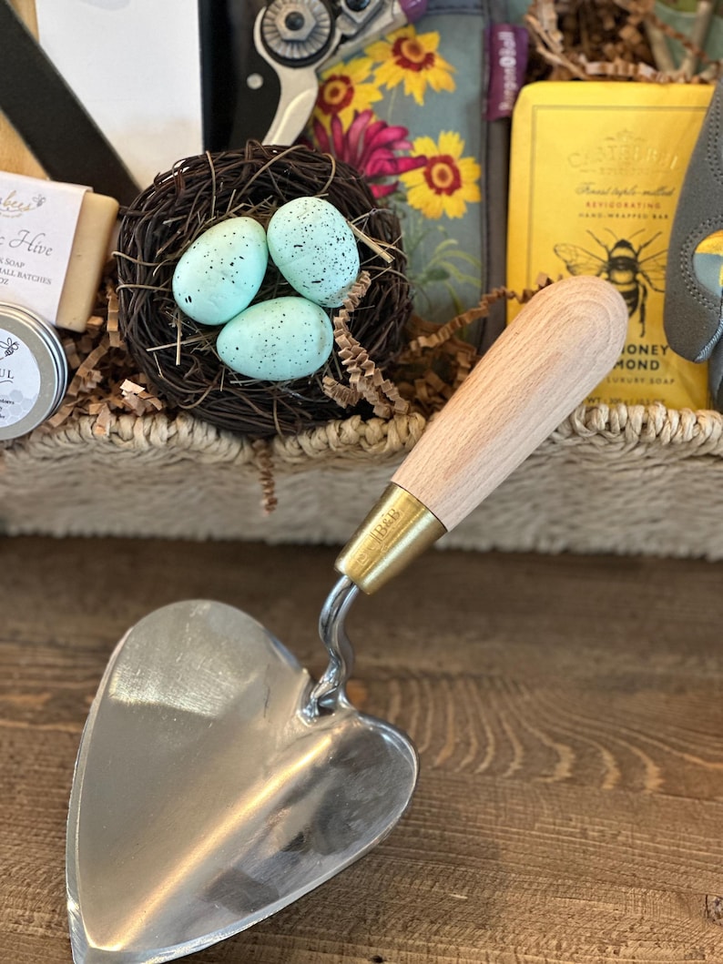 May include: A heart-shaped gardening trowel with a wooden handle and a brass ferrule. The trowel is sitting on a wooden surface.