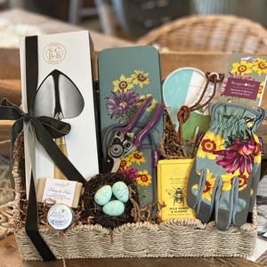 May include: A wicker basket filled with gardening themed gifts. The basket includes a pair of gardening gloves, a trowel, a pair of pruning shears, a tin with a floral design, a small jar of honey, a bar of soap, and a small tin of hand cream. The basket is decorated with a black ribbon and a nest with three blue eggs.