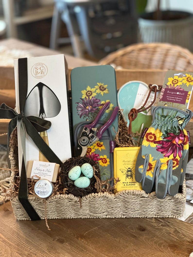 May include: A wicker basket filled with gardening themed gifts. The basket includes a pair of gardening gloves, a trowel, a pair of pruning shears, a tin with a floral design, a small jar of honey, a bar of soap, and a small tin of hand cream. The basket is decorated with a black ribbon and a nest with three blue eggs.