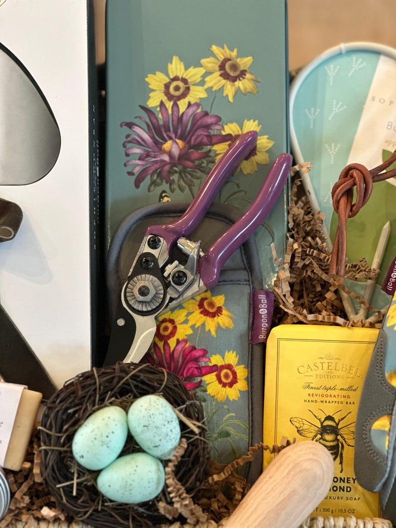 May include: A gardening gift basket with a pair of purple pruning shears, a bar of soap, a wooden spoon, and a bird's nest with blue eggs. The basket is filled with brown paper shreds and has a floral pattern.