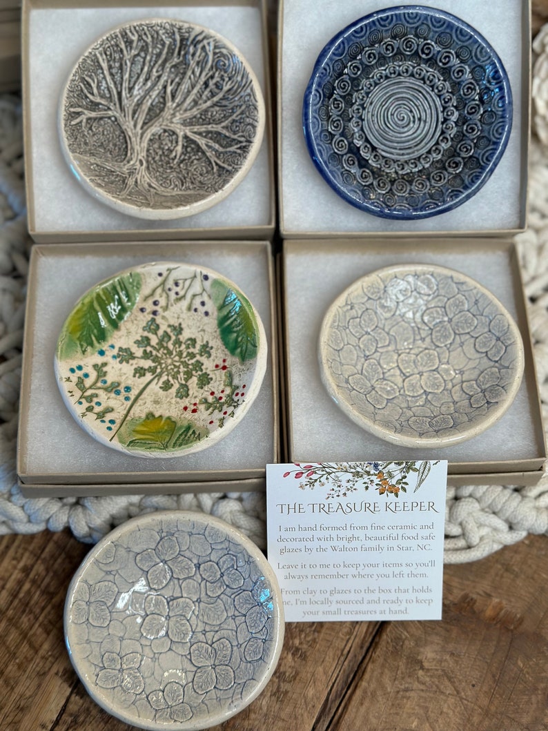 May include: Four small ceramic dishes with different designs. The dishes are in white boxes with a white background. The dishes are all about 4 inches in diameter. The designs are a tree, a swirl pattern, a floral pattern, and a textured pattern. The text on the card reads "The Treasure Keeper. I am hand formed from fine ceramic and decorated with bright, beautiful food safe glazes by the Walton family in Star, NC. Leave it to me to keep your items so you'll always remember where you left them. From clay to glazes to the box that holds me, I'm locally sourced and ready to keep your small treasures at hand."