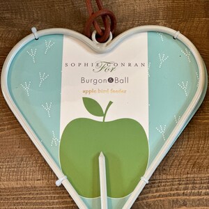 May include: A white heart-shaped bird feeder with a green apple design. The feeder is made by Sophie Conran for Burgon & Ball. The text "apple bird feeder" is printed on the feeder.