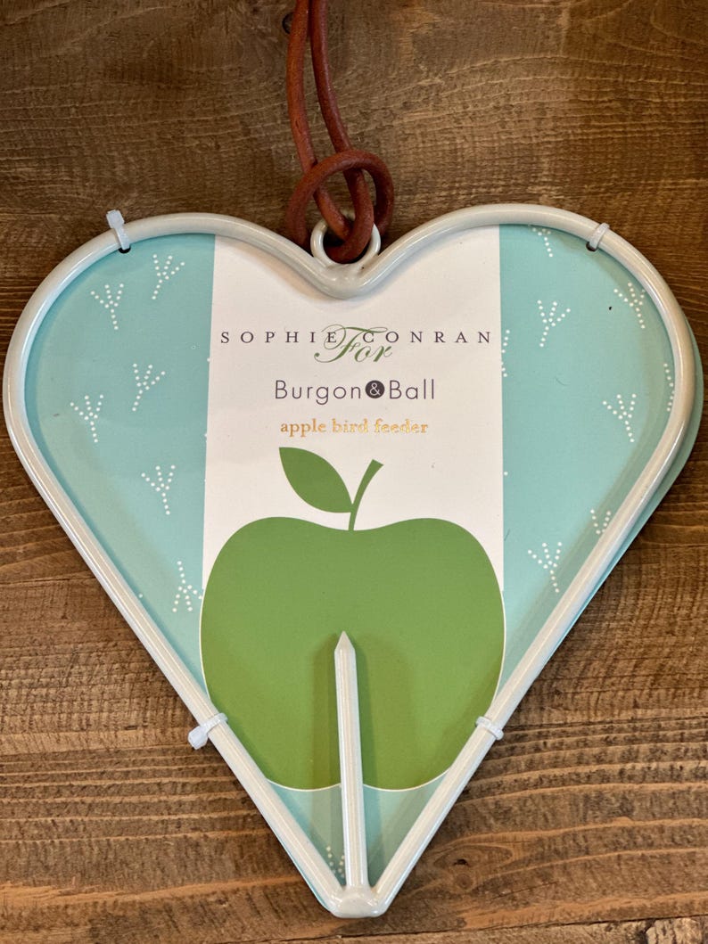 May include: A white heart-shaped bird feeder with a green apple design. The feeder is made by Sophie Conran for Burgon & Ball. The text "apple bird feeder" is printed on the feeder.