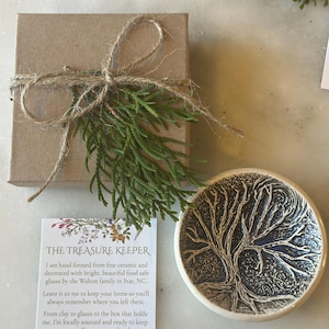 May include: A small, white ceramic dish with a blue tree design. The dish is hand-crafted and food safe. The dish is part of a set of handmade pottery items. The text on the card reads: "THE TREASURE KEEPER I am hand formed from fine ceramic and decorated with bright, beautiful food safe glazes by the Walton family in Star, NC. Leave it to me to keep your items so you'll always remember where you left them. From clay to glazes to the box that holds me, I'm locally sourced and ready to keep your small treasures at hand."
