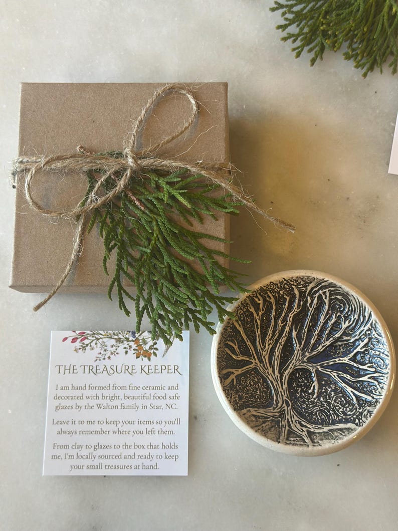May include: A small, white ceramic dish with a blue tree design. The dish is hand-crafted and food safe. The dish is part of a set of handmade pottery items. The text on the card reads: "THE TREASURE KEEPER I am hand formed from fine ceramic and decorated with bright, beautiful food safe glazes by the Walton family in Star, NC. Leave it to me to keep your items so you'll always remember where you left them. From clay to glazes to the box that holds me, I'm locally sourced and ready to keep your small treasures at hand."