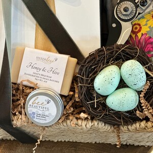 May include: A wicker basket filled with a variety of items, including a bar of goat's milk soap labeled "Honey & Hive", a small tin of hand cream labeled "bee BEAUTIFUL", and a nest with three blue speckled eggs.
