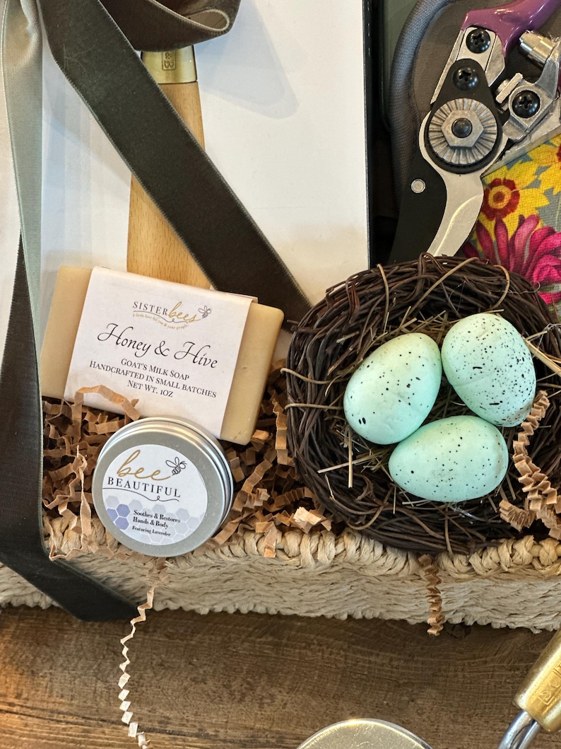 May include: A wicker basket filled with a variety of items, including a bar of goat's milk soap labeled "Honey & Hive", a small tin of hand cream labeled "bee BEAUTIFUL", and a nest with three blue speckled eggs.