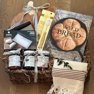 May include: A bread-making gift hamper featuring a wooden chopping board, a cookbook titled "Wild Bread", and baking utensils. Includes jars of preserves, a striped towel, and a decorative ribbon.