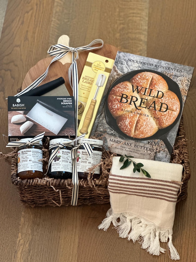 May include: A bread-themed gift basket with a wooden cutting board, a cookbook titled "Wild Bread", and various baking tools. Includes jars of preserves, a striped towel, and a decorative ribbon.