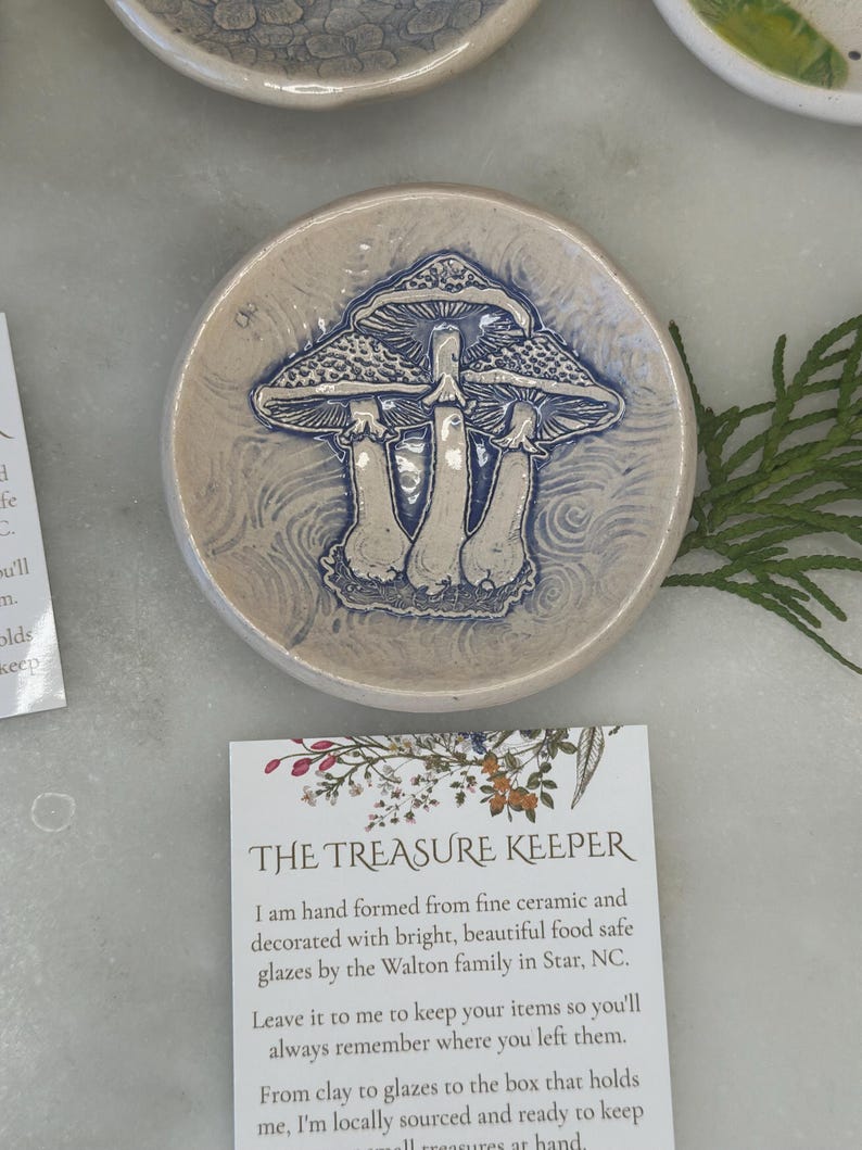 May include: A small, white ceramic dish with a blue and white mushroom design. The dish is hand-formed and decorated with food-safe glazes. The text "THE TREASURE KEEPER" is printed on a card next to the dish. The card reads: "I am hand formed from fine ceramic and decorated with bright, beautiful food safe glazes by the Walton family in Star, NC. Leave it to me to keep your items so you'll always remember where you left them. From clay to glazes to the box that holds me, I'm locally sourced and ready to keep all treasures at hand."