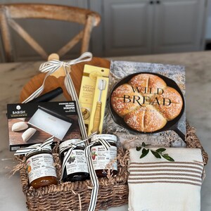 May include: A bread-making gift basket with a wooden cutting board, a cookbook titled "Wild Bread", a bench scraper, a bread lame, and jars of preserves. The basket includes a striped towel and is tied with a ribbon.