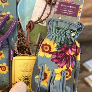 May include: A gift basket filled with gardening supplies, including a pair of gray gardening gloves with a floral print, a bar of soap, and a small wooden tool. The gloves have the text "Burgon & Ball" on the wrist.