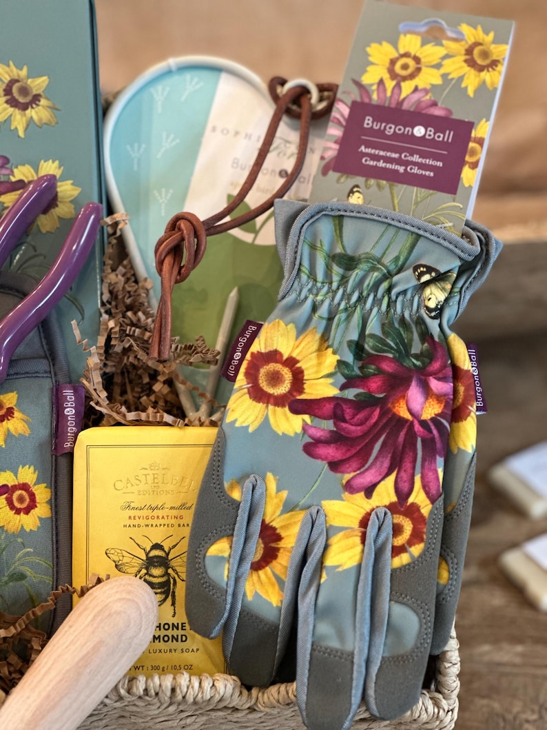 May include: A gift basket filled with gardening supplies, including a pair of gray gardening gloves with a floral print, a bar of soap, and a small wooden tool. The gloves have the text "Burgon & Ball" on the wrist.