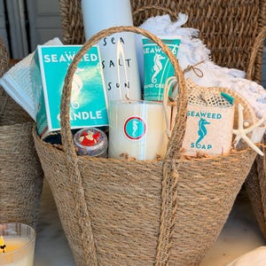 May include: A woven basket filled with spa products. The basket contains a white tumbler with "Sea Salt Sun" text, a candle, a box of seaweed candle, a tube of hand cream, and a bar of seaweed soap. A starfish and a white towel are also in the basket.