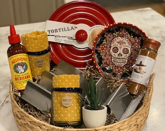 May include: A wicker basket filled with a variety of Mexican-themed food and cooking items, including a red tortilla warmer, a small plant, a bottle of hot sauce, a jar of spices, and a small plate with a sugar skull design.
