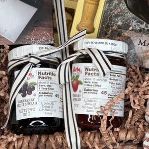 May include: Two jars of fruit spread, one blueberry and one strawberry, are tied with a striped ribbon. The jars are in a woven basket with shredded paper. Each jar has a label with nutrition facts and the brand name.
