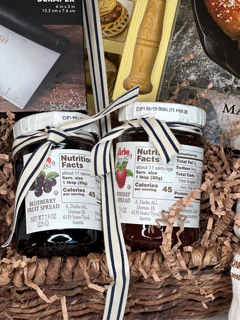May include: Two jars of fruit spread, one blueberry and one strawberry, are tied with a striped ribbon. The jars are in a woven basket with shredded paper. Each jar has a label with nutrition facts and the brand name.