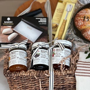 May include: A gift basket filled with gourmet food items. It includes jars of fruit spread, a bench scraper, a bread lame, and a wooden cutting board. The basket is decorated with a striped ribbon.