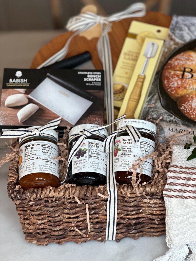 May include: A gift basket filled with gourmet food items. It includes jars of fruit spread, a bench scraper, a bread lame, and a wooden cutting board. The basket is decorated with a striped ribbon.