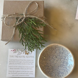 May include: A small, white ceramic dish with a speckled glaze pattern. The dish is hand-formed and food safe. The dish is packaged in a brown box with twine and a sprig of greenery. The text on the card reads "The Treasure Keeper. I am hand formed from fine ceramic and decorated with bright, beautiful food safe glazes by the Walton family in Star, NC. Leave it to me to keep your items so you'll always remember where you left them. From clay to glazes to the box that holds me, I'm locally sourced and ready to keep your small treasures at hand."