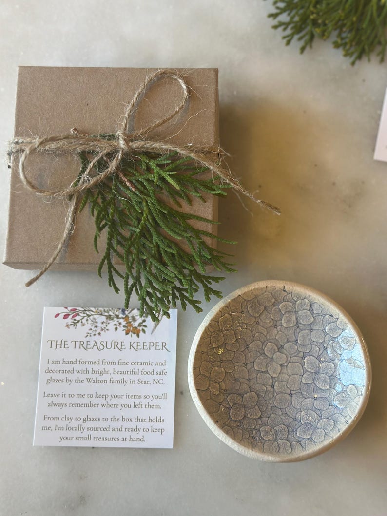 May include: A small, white ceramic dish with a speckled glaze pattern. The dish is hand-formed and food safe. The dish is packaged in a brown box with twine and a sprig of greenery. The text on the card reads "The Treasure Keeper. I am hand formed from fine ceramic and decorated with bright, beautiful food safe glazes by the Walton family in Star, NC. Leave it to me to keep your items so you'll always remember where you left them. From clay to glazes to the box that holds me, I'm locally sourced and ready to keep your small treasures at hand."