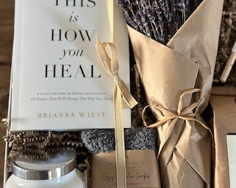 May include: A white gift box filled with a self-help book titled "This is How You Heal" by Brianna Wiest, a bouquet of lavender, a gray cozy water sock, and a white candle with a silver lid.